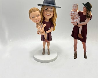May include: Figurines of a woman holding a child, and another woman holding a child. The figurines are wearing burgundy dresses and hats. The child figurines are wearing plaid shirts. The figurines are on a white surface.