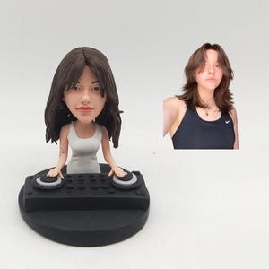 May include: A figurine of a person with dark hair and a white dress, posed as a DJ behind a black console. The figurine has a caricature-like appearance. A photo of a person with similar hair and a black vest top is next to the figurine.