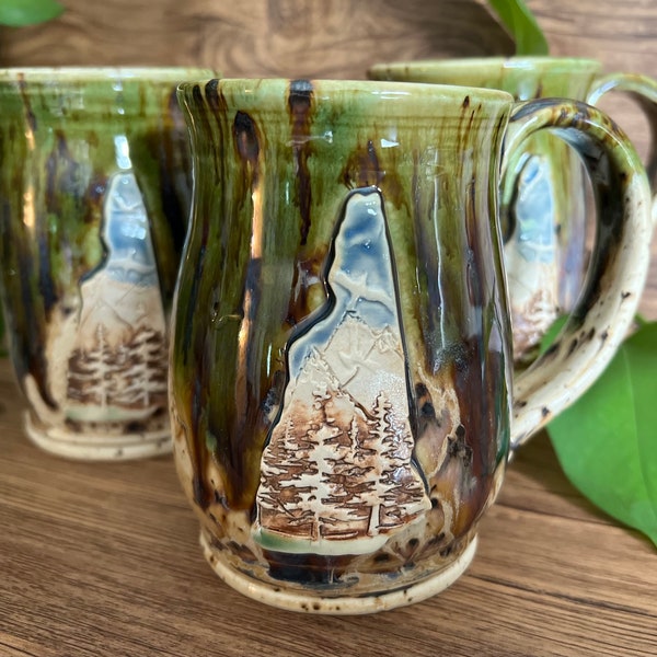 May include: Three ceramic mugs with a green, brown and white glaze. Each mug features a raised design of a mountain scene with trees.