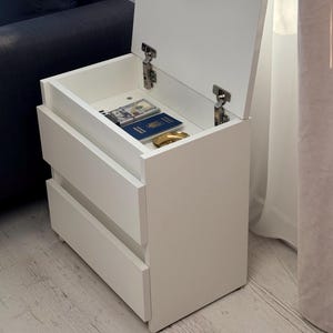 Pode incluir: Uma mesa de cabeceira branca com duas gavetas e uma tampa articulada. A tampa está aberta, revelando um compartimento com um passaporte, dinheiro e um pequeno objeto dourado. A mesa de cabeceira está ao lado de uma janela com cortinas transparentes.