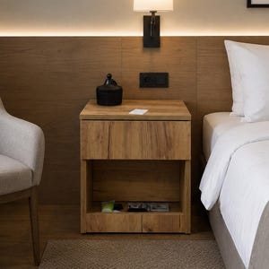 May include: A hotel bedroom interior with a wooden bedside table, a lamp, and a framed picture. The bed is made with white bedding and a grey throw. A grey armchair and a small table with a mug are also in the scene.