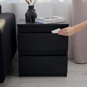 Pode incluir: Uma mesa de cabeceira preta com duas gavetas. Um vaso com plantas secas e um livro intitulado "Delicious Places" estão em cima. Uma mão está a alcançar a gaveta superior. A mesa de cabeceira está ao lado de um sofá preto.