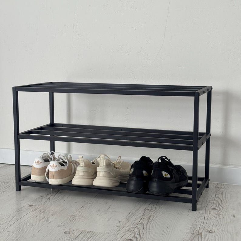 Op de afbeelding: Een zwart metalen schoenenrek met drie niveaus, met verschillende paren schoenen. De schoenen zijn onder meer witte, beige en zwarte sneakers. Het rek staat tegen een witte muur en staat op een lichtgekleurde houten vloer.