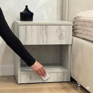 Könnte beinhalten: Ein Nachttisch aus hellgrauem Holz mit einer Schublade und einem offenen Regal. Ein schwarzes Deko-Objekt steht obenauf. Der Nachttisch steht neben einem Bett mit beigem Kopfteil und einem Stapel Decken. Das Holz hat ein hellgraues Maserungsmuster.