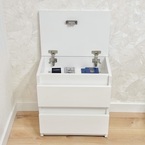 May include: A white storage cabinet with a hinged lid and two drawers. The lid is open, showing a small compartment containing a few items. The cabinet is positioned against a patterned wall and a wooden floor.