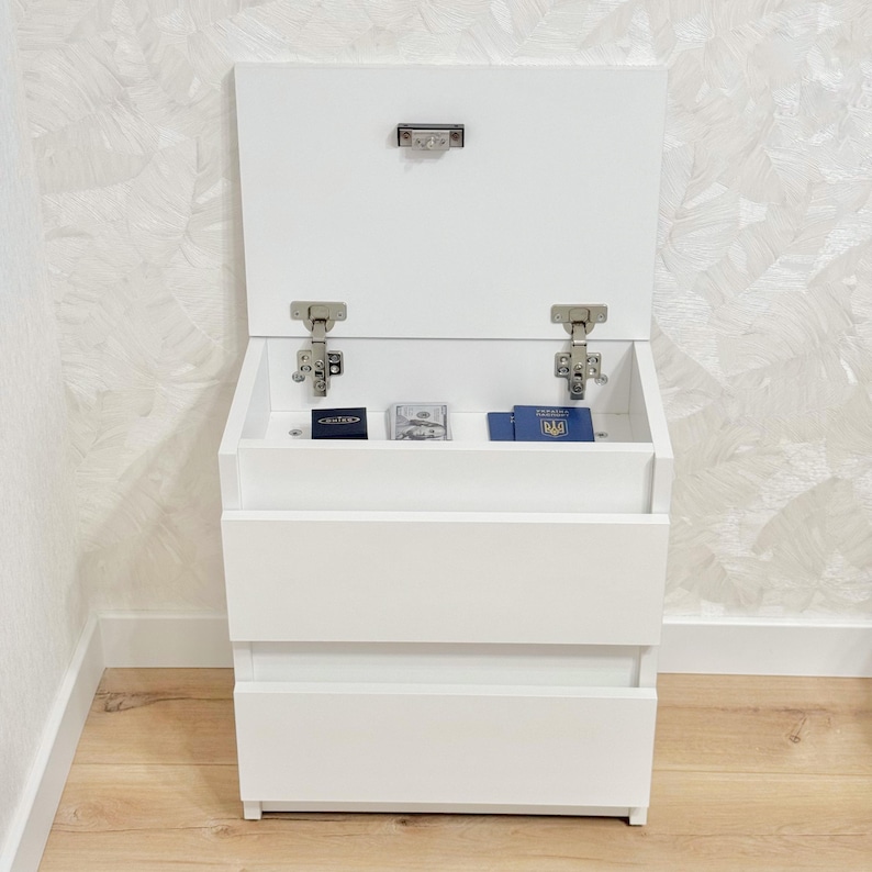 May include: A white storage cabinet with a hinged lid and two drawers. The lid is open, showing a small compartment containing a few items. The cabinet is positioned against a patterned wall and a wooden floor.