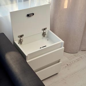 May include: A white bedside cabinet with two drawers and a lift-up top. The top is open, showing the inside. The cabinet has silver hinges and a latch. It is positioned next to a dark grey upholstered sofa.