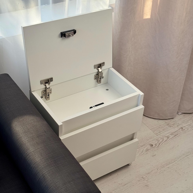 May include: A white bedside cabinet with two drawers and a lift-up top. The top is open, showing the inside. The cabinet has silver hinges and a latch. It is positioned next to a dark grey upholstered sofa.