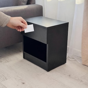 Pode incluir: Uma mesa de cabeceira preta com um design retangular. A mesa de cabeceira tem uma superfície superior e uma prateleira aberta. Uma mão segura um cartão branco perto da parte superior da mesa de cabeceira. A mesa está em um piso de madeira clara.