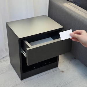 May include: A dark gray nightstand with a single drawer open, revealing a white interior. The nightstand is rectangular and sits on a light gray floor. A hand holds a white card near the open drawer.
