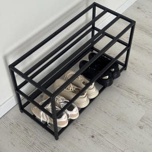 Op de afbeelding: Een zwart metalen schoenenrek met drie niveaus, met verschillende paren schoenen. De schoenen zijn in verschillende kleuren, waaronder zwart, beige en wit. Het rek is rechthoekig en heeft een minimalistisch ontwerp.