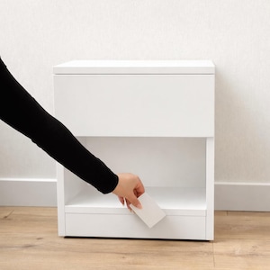 May include: A white bedside table with a drawer and an open shelf. A hand is reaching into the open shelf to place a small white card. The nightstand is made of wood and has a minimalist design.