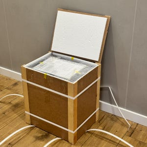 May include: A square wooden shipping crate with a hinged lid held open by a white foam panel. The crate is reinforced with light-coloured wooden corner supports and secured by white plastic strapping bands. Inside, a wrapped item is visible.