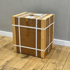 May include: A large, square cardboard box with wooden supports and white straps. The box is brown with a few stickers and is sitting on a wooden floor. The box is ready for shipping.