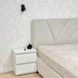 May include: A white two-drawer bedside table stands next to a bed with a beige upholstered headboard featuring geometric paneling and a textured beige bedspread. A small black object rests on the bedside table surface.