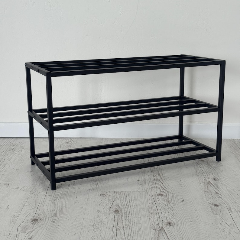 Op de afbeelding: Een zwart metalen schoenenrek met drie niveaus. Het rek heeft een rechthoekig frame en horizontale staven voor schoenenopslag. Het ontwerp is eenvoudig en modern, geschikt voor entrees of kasten.