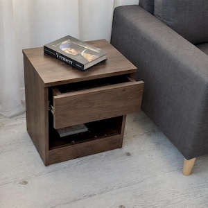 May include: A brown wooden nightstand with a drawer slightly open, revealing the interior. A book titled "Vermeer" rests on top. The nightstand is next to a gray sofa. The nightstand measures approximately 20 inches tall, 16 inches wide, and 14 inches deep.