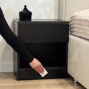 Può includere: Un comodino nero con un cassetto e un ripiano aperto. Una scatola nera decorativa con un nappa è sopra. Il comodino è accanto a un letto con una testiera beige e coperte piegate.