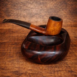 May include: A wooden tobacco pipe rests on a dark brown, rounded ashtray. The pipe has a light brown bowl and a dark brown stem. The ashtray is made of a darker wood with visible grain patterns. The background is a wooden surface.
