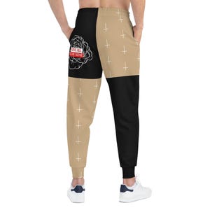May include: Black and tan sweatpants with a split design. One side features a black panel with a white rose graphic and the text "LOVE ALL FEAR NONE". The other side is tan with white crosses.