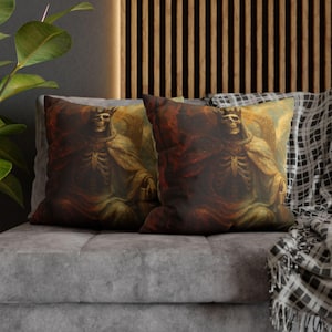 May include: Two cushions with a gothic artwork of a crowned skeleton in a robe. The design uses a palette of browns, reds, and golds, giving a vintage feel. The cushions are placed on a grey sofa, with a patterned throw.