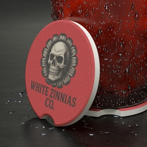May include: A red car coaster with a white border and a skull design inside a flower. The text "WHITE ZINNIAS CO." is printed below the skull. The coaster is next to a glass with condensation.