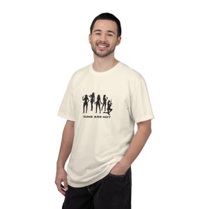 May include: An off-white short-sleeved t-shirt featuring a black graphic print of five silhouettes of individuals holding firearms. Below the silhouettes, the text "GUNS ARE HOT" is printed in a bold, black, sans-serif font on the chest area.