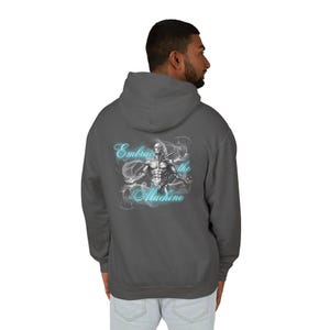 May include: A charcoal gray hoodie with a hood. The back of the hoodie features a detailed illustration of a cyborg with the words "Embrace the Machine" in a light blue, stylized font. The cyborg is surrounded by a smoky effect.