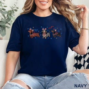 May include: Navy blue t-shirt featuring three dachshunds with patriotic bow ties and hats. The dogs are brown, gray, and black, with red, white, and blue accents. Blue stars are scattered around the dogs.