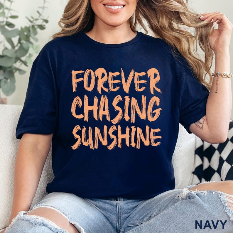 May include: A navy blue short-sleeved t-shirt featuring the text "FOREVER CHASING SUNSHINE" printed in a light orange, brush-stroke style font across the chest. The shirt has a crew neckline and a standard fit, with the word "NAVY" in the corner.