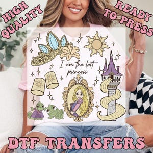 May include: A light pink t-shirt featuring DTF transfers of a princess theme. The design includes a crown, sun, tower, and the text "I am the lost princess". Additional graphics include a lily, lanterns, and a princess in a frame.