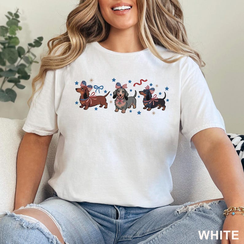 May include: White t-shirt featuring three dachshunds with patriotic bows and star accents. The dogs are brown, gray, and black, with red, white, and blue details. The word "WHITE" is printed at the bottom.