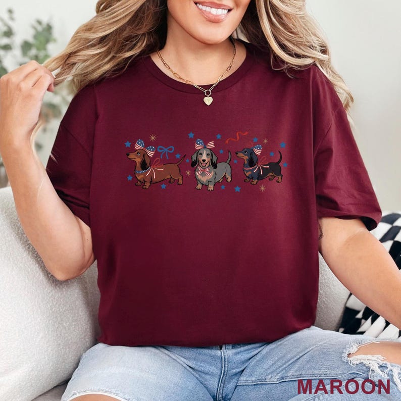 May include: Maroon t-shirt featuring three dachshunds with patriotic bows and flags. The dogs are brown, gray, and black, with blue stars and red and blue ribbons. The word "MAROON" is printed at the bottom.