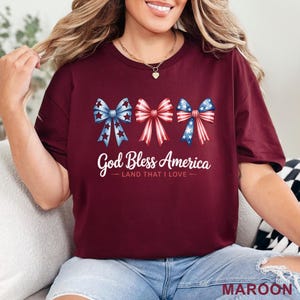 May include: Maroon t-shirt featuring three patriotic bows in red, white, and blue. The text "God Bless America - LAND THAT I LOVE" is printed below the bows. The word "MAROON" is printed at the bottom of the shirt.