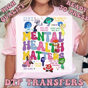 May include: A pink t-shirt featuring the phrase "MENTAL HEALTH MATTERS" in colorful bubble letters, surrounded by cartoon characters and positive affirmations. The design includes the text "DTF TRANSFERS" and "HIGH QUALITY READY TO PRESS".