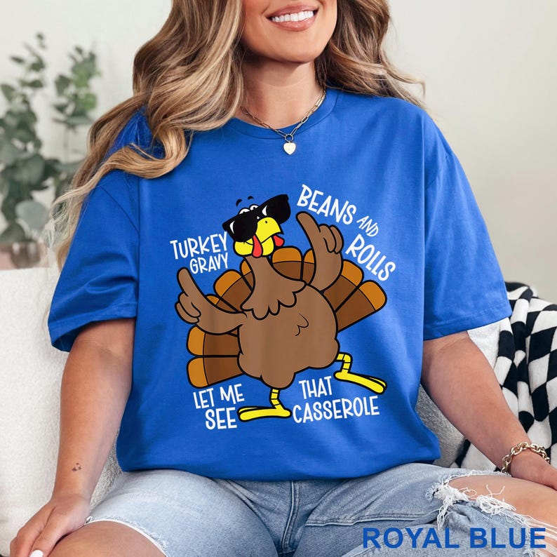 Puede incluir: Camiseta azul real con un pavo de dibujos animados con gafas de sol y gesticulando con las alas. El texto de la camiseta dice: "Turkey Gravy, Beans and Rolls, Let Me See That Casserole."