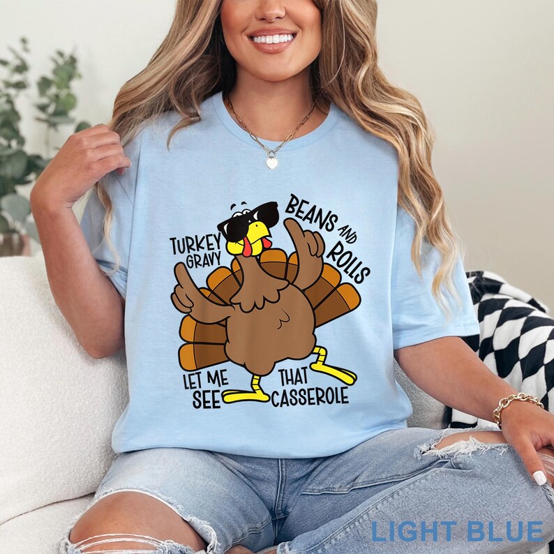 Puede incluir: Camiseta azul claro con un pavo de dibujos animados con gafas de sol y gesticulando con las alas. El texto en la camiseta incluye "Turkey Gravy", "Beans and Rolls" y "Let Me See That Casserole".