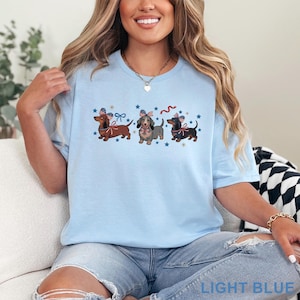 May include: Light blue t-shirt featuring three dachshunds with patriotic bows and accents. The dogs are brown, gray, and black, with blue stars and red and blue ribbons. The text "LIGHT BLUE" is at the bottom of the shirt.