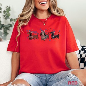 May include: Red t-shirt featuring three dachshunds with patriotic bows and stars. The dogs are in brown, gray, and black, with red, white, and blue accents. The word "RED" is printed in the bottom right corner.