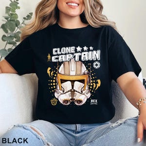 May include: Black t-shirt featuring a graphic of a Clone Captain helmet with the text "CLONE CAPTAIN". The design includes stars, a symbol, and the name "REX". The shirt is a crew neck style.