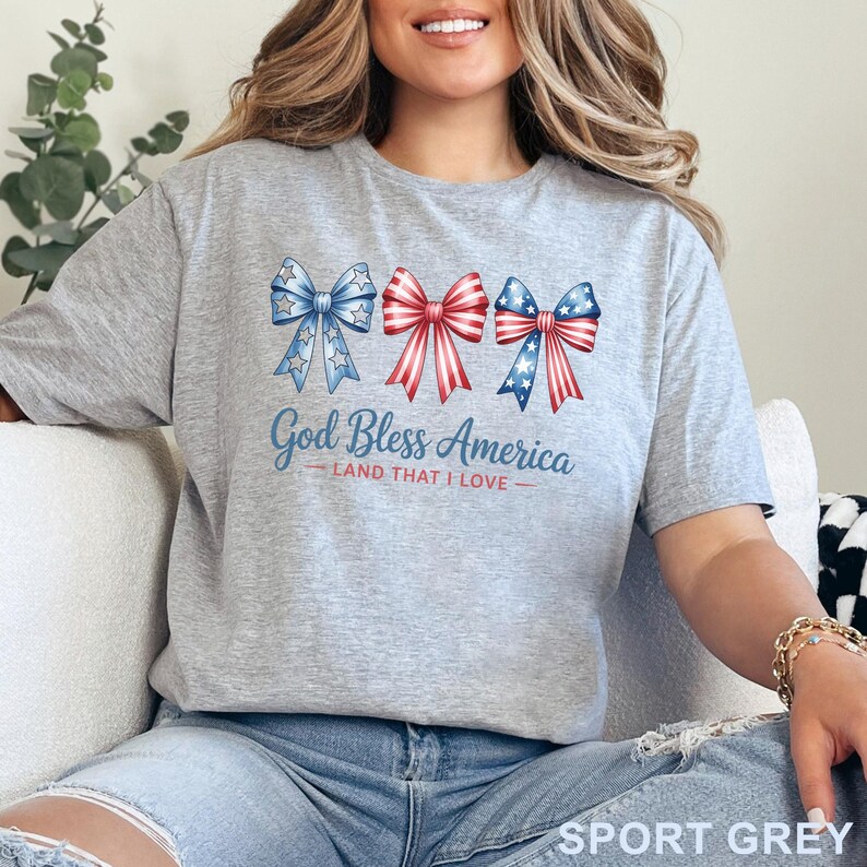 May include: Sport grey t-shirt with the text "God Bless America - Land That I Love." The shirt features three bows in red, white, and blue, with star and stripe patterns. A patriotic design for the USA.