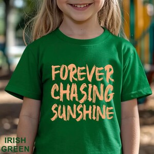 May include: A short-sleeved Irish green t-shirt featuring the text FOREVER CHASING SUNSHINE printed in a light peach, brush-stroke style font across the centre of the chest. The garment has a crew neckline and a standard straight hem.