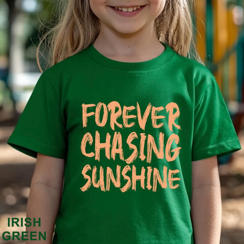 May include: A short-sleeved Irish green t-shirt featuring the text FOREVER CHASING SUNSHINE printed in a light peach, brush-stroke style font across the centre of the chest. The garment has a crew neckline and a standard straight hem.