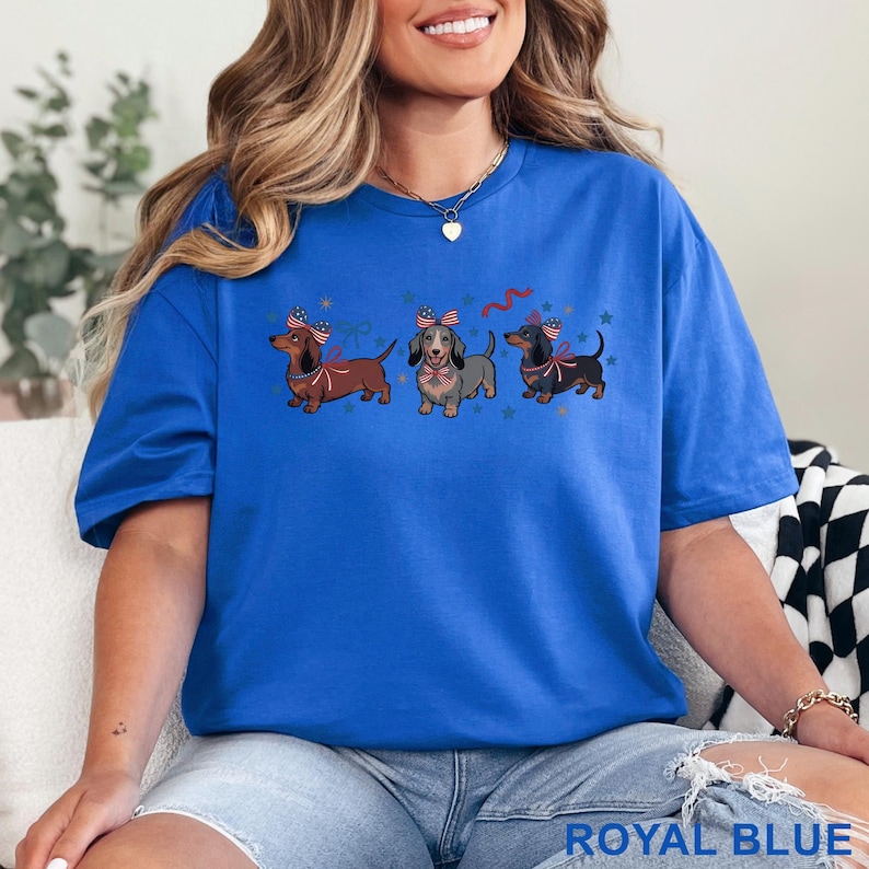 May include: Royal blue t-shirt featuring three dachshunds with patriotic bows and star accents. The shirt's design includes red, white, and blue elements, perfect for celebrating a national holiday. The text "ROYAL BLUE" is printed at the bottom of the shirt.