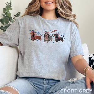 May include: A light gray t-shirt featuring three dachshunds with patriotic bows and stars. The text "SPORT GREY" is printed at the bottom of the shirt. The dachshunds are brown, gray, and black.