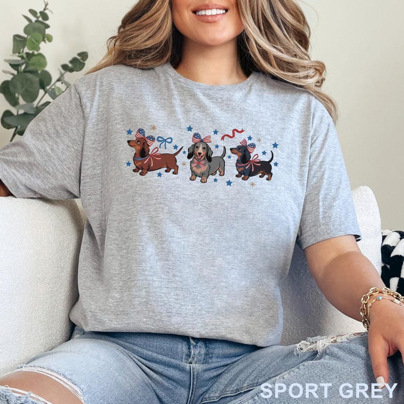 May include: A light gray t-shirt featuring three dachshunds with patriotic bows and stars. The text "SPORT GREY" is printed at the bottom of the shirt. The dachshunds are brown, gray, and black.