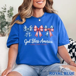 May include: Royal blue t-shirt featuring three patriotic bow designs in red, white, and blue. The text "God Bless America - LAND THAT I LOVE" is printed below the bows. The shirt is a comfortable, casual style.