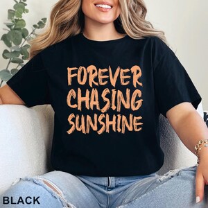 May include: A black short-sleeved t-shirt featuring the text "FOREVER CHASING SUNSHINE" printed in a light orange, brush-stroke style font across the chest. The shirt has a crew neckline and a standard fit, with the word "BLACK" printed in the corner.