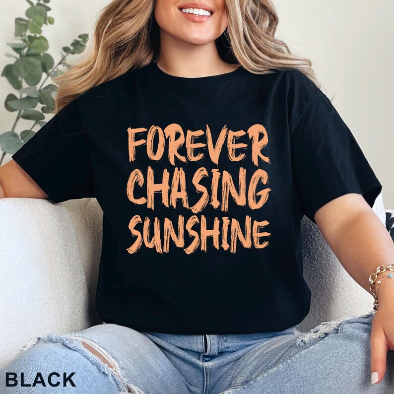 May include: A black short-sleeved t-shirt featuring the text "FOREVER CHASING SUNSHINE" printed in a light orange, brush-stroke style font across the chest. The shirt has a crew neckline and a standard fit, with the word "BLACK" printed in the corner.