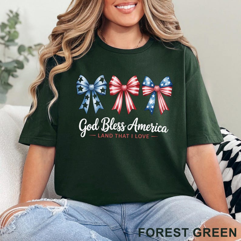 May include: Forest green t-shirt featuring three patriotic bow designs in red, white, and blue. The text "God Bless America - Land That I Love" is printed below the bows. The shirt is a comfortable, casual style.